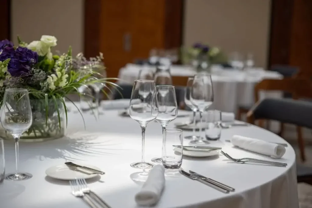 A round dining table set for a formal event with white tablecloth, glassware, plates, cutlery, and a floral centerpiece. Other similarly set tables are visible in the background.